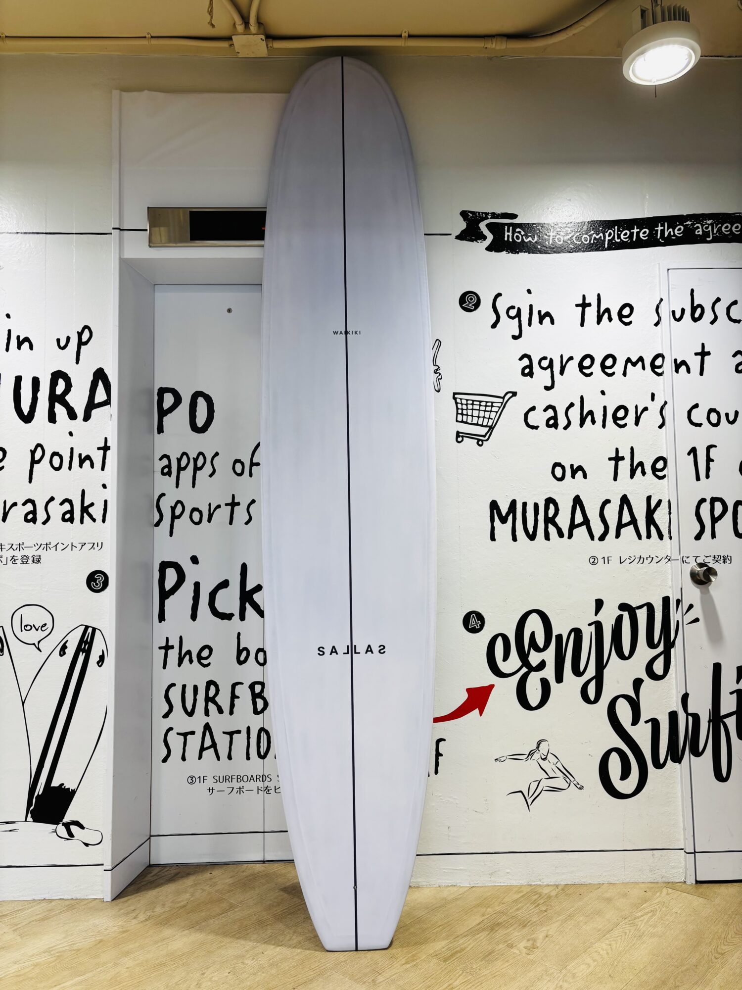 IMG_0469 - SHOP NEWS | ムラサキスポーツオンラインストア 通販 White surfboard standing upright between walls covered with bold black handwritten text and small drawings, on a wooden floor.