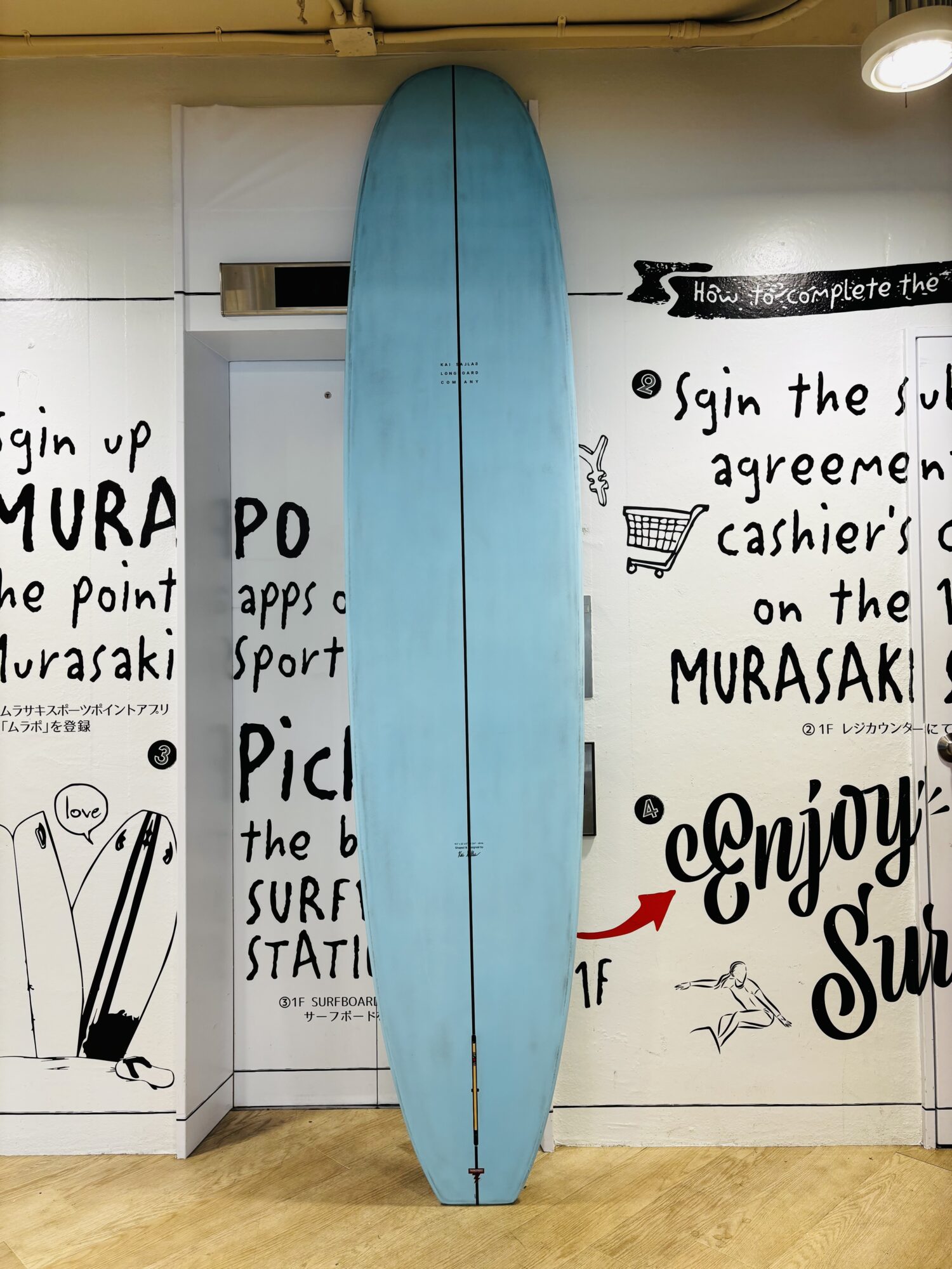 IMG_0481 - SHOP NEWS | ムラサキスポーツオンラインストア 通販 Blue surfboard stands upright against a white wall covered in black handwritten text and small drawings in a shop interior.
