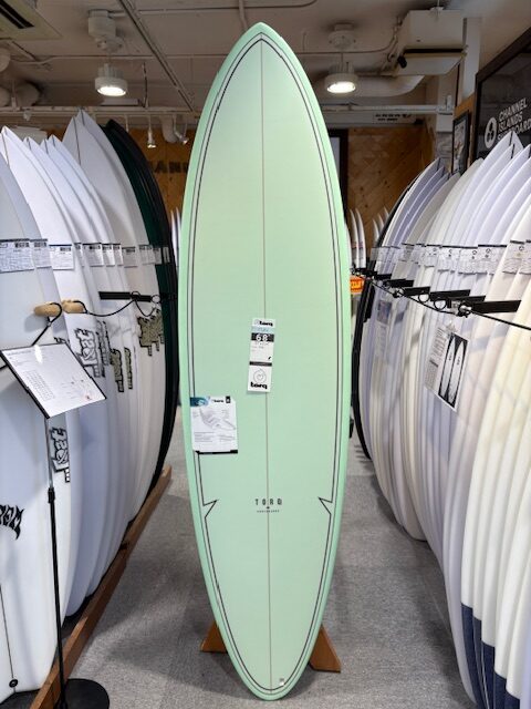 Mint-green surfboard standing upright on a showroom display, with price tags attached and rows of white boards on either side in a shop.