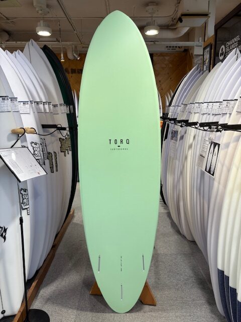 Mint green longboard surfboard on a display stand in a surf shop, surrounded by white boards on racks, center brand reads TORO Surfboards