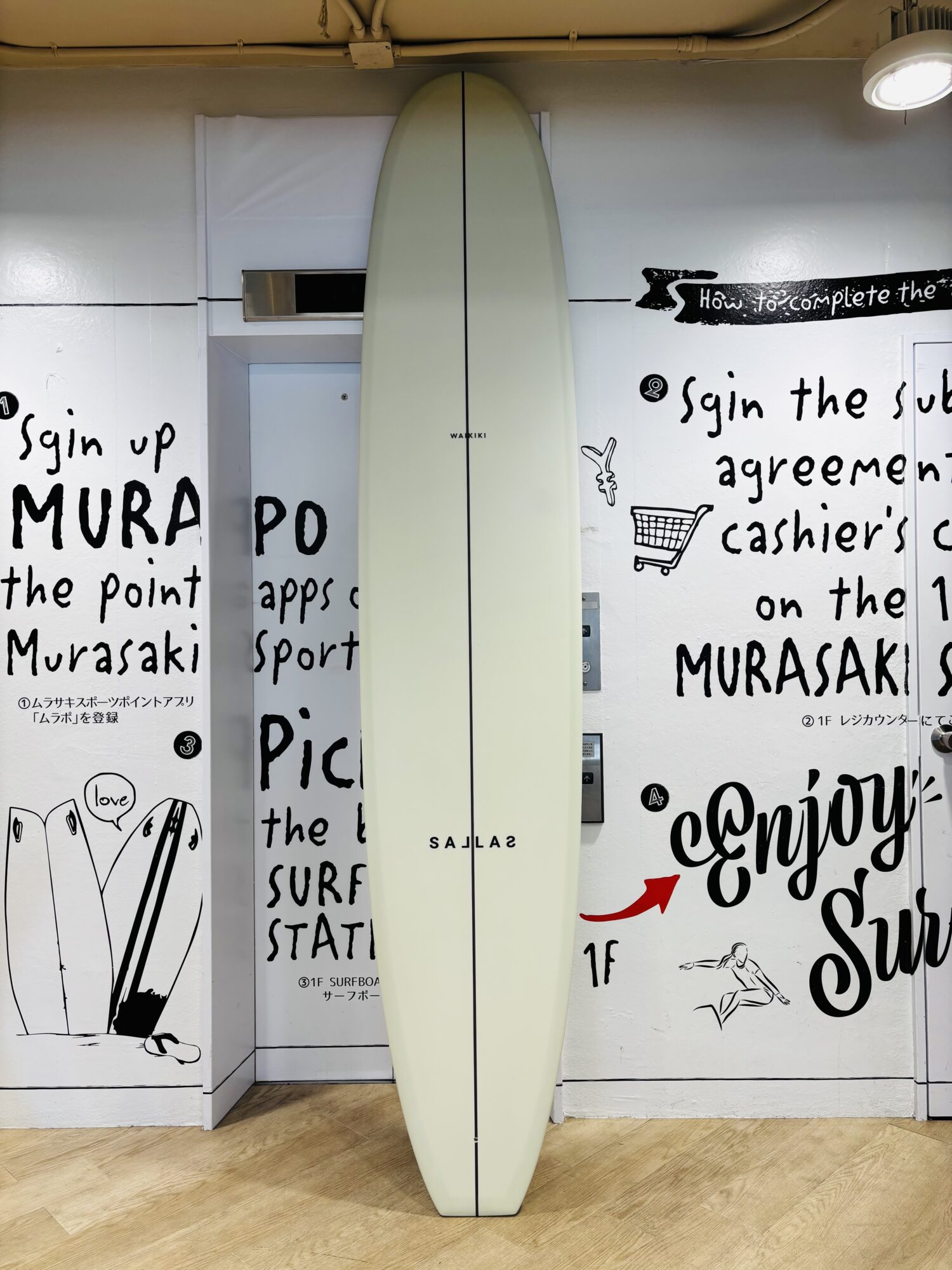 IMG_8973 - SHOP NEWS | ムラサキスポーツオンラインストア 通販 Off-white surfboard standing upright indoors against a wall with black graffiti-style text and illustrations.