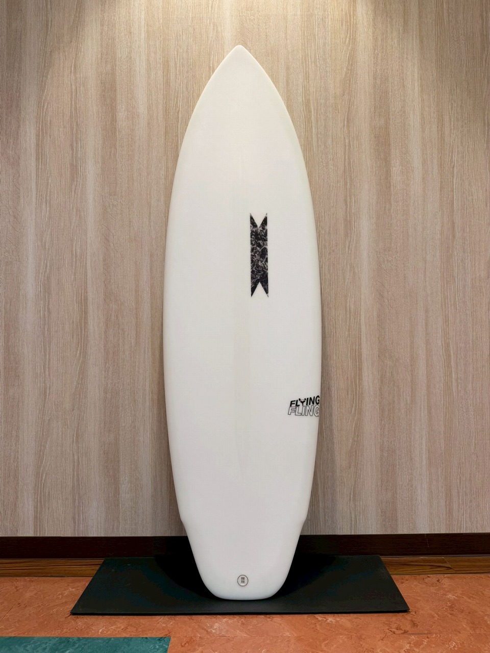 LINE_ALBUM_2026 SB_260530_1 - SHOP NEWS | ムラサキスポーツオンラインストア 通販 White surfboard standing upright on a black mat against a wood-paneled wall, with a small black central logo and a round tail plug.