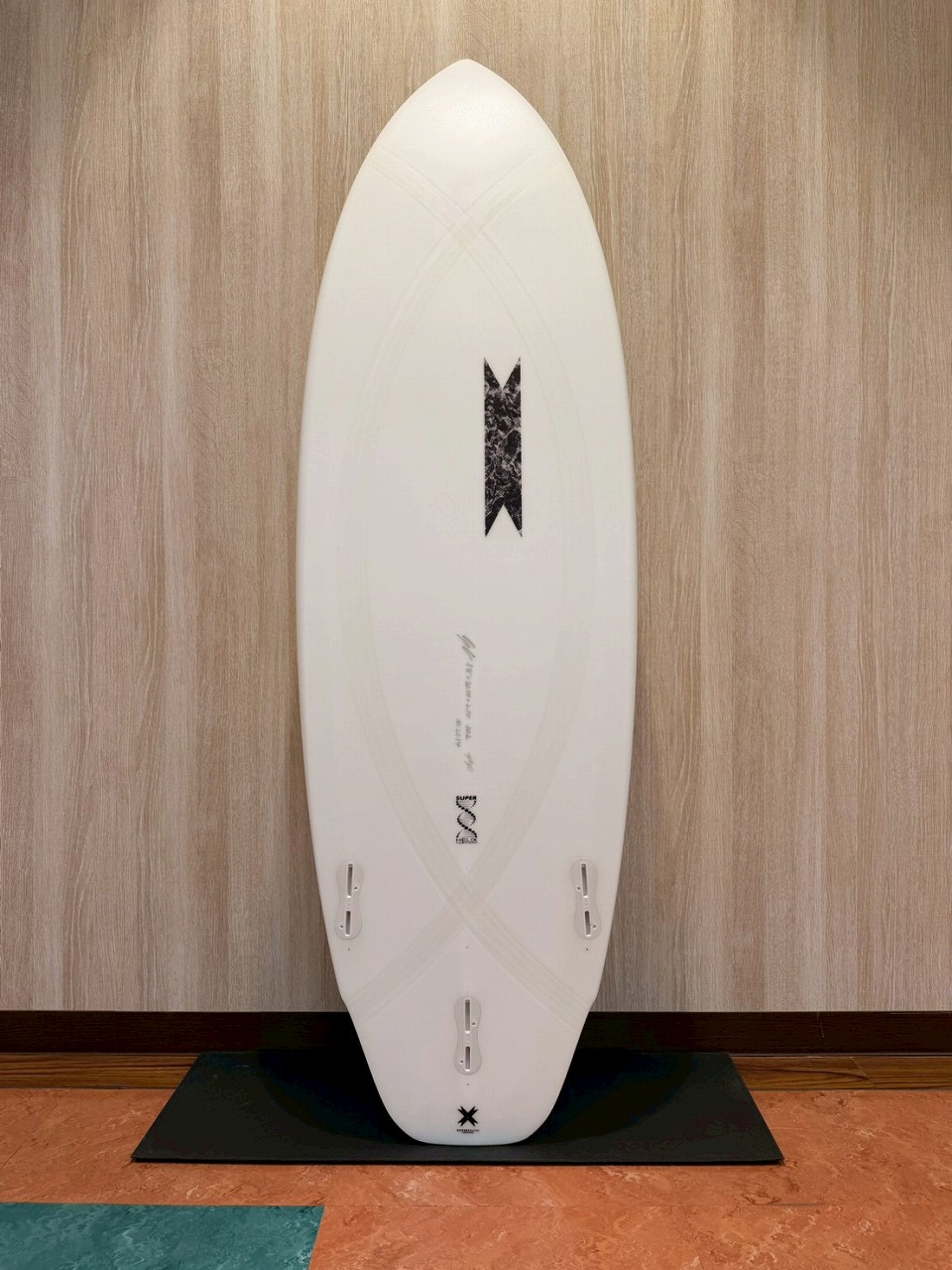LINE_ALBUM_2026 SB_260530_2 - SHOP NEWS | ムラサキスポーツオンラインストア 通販 White surfboard standing upright on a black mat against a wood-paneled wall; three fin boxes visible near the bottom.