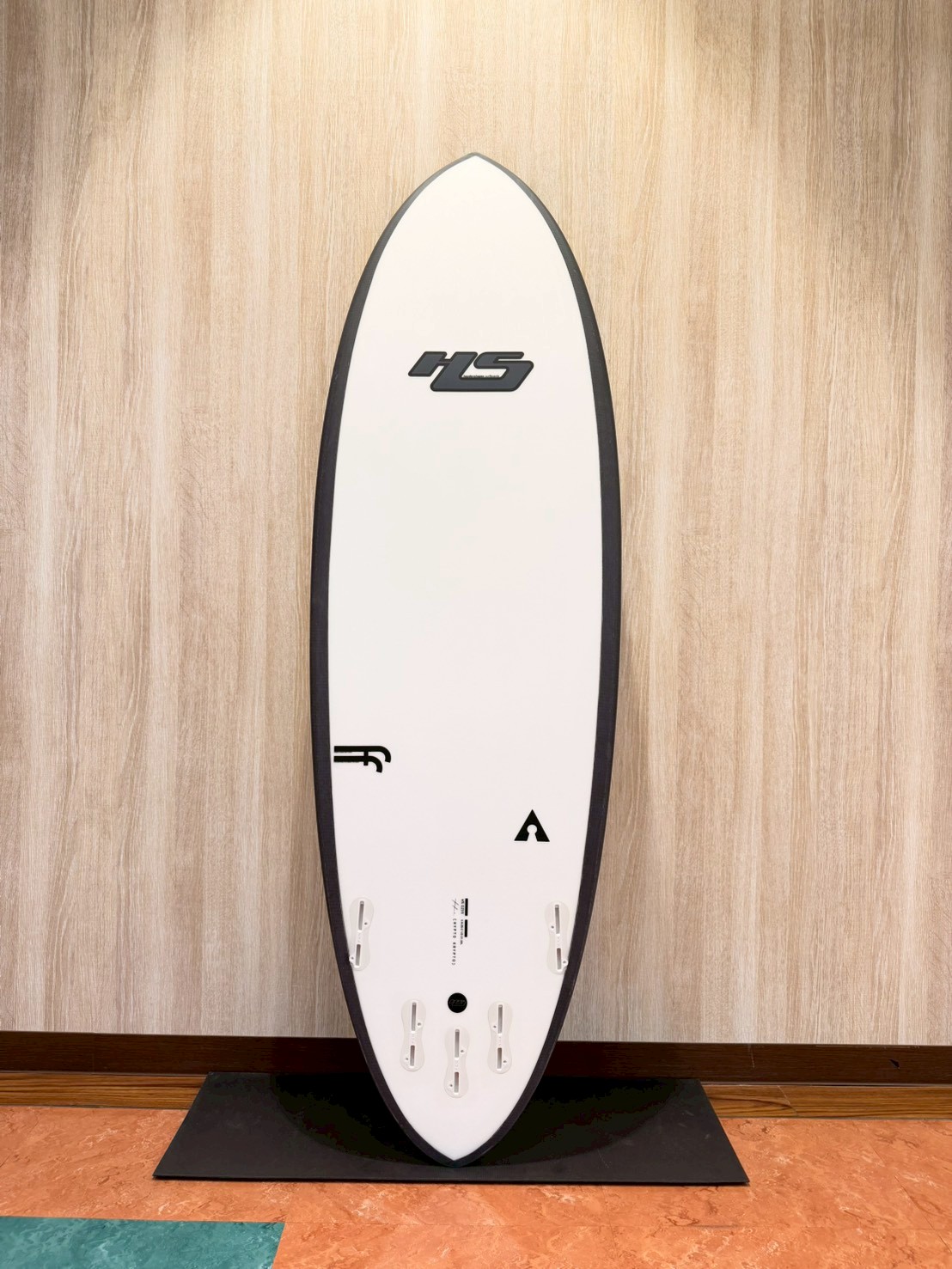 LINE_ALBUM_2026 ヘイデン_260427_1 - SHOP NEWS | ムラサキスポーツオンラインストア 通販 White surfboard with black rails standing upright on a black display mat against a wood-paneled wall, logo near the center and fin/foot well slots at the bottom