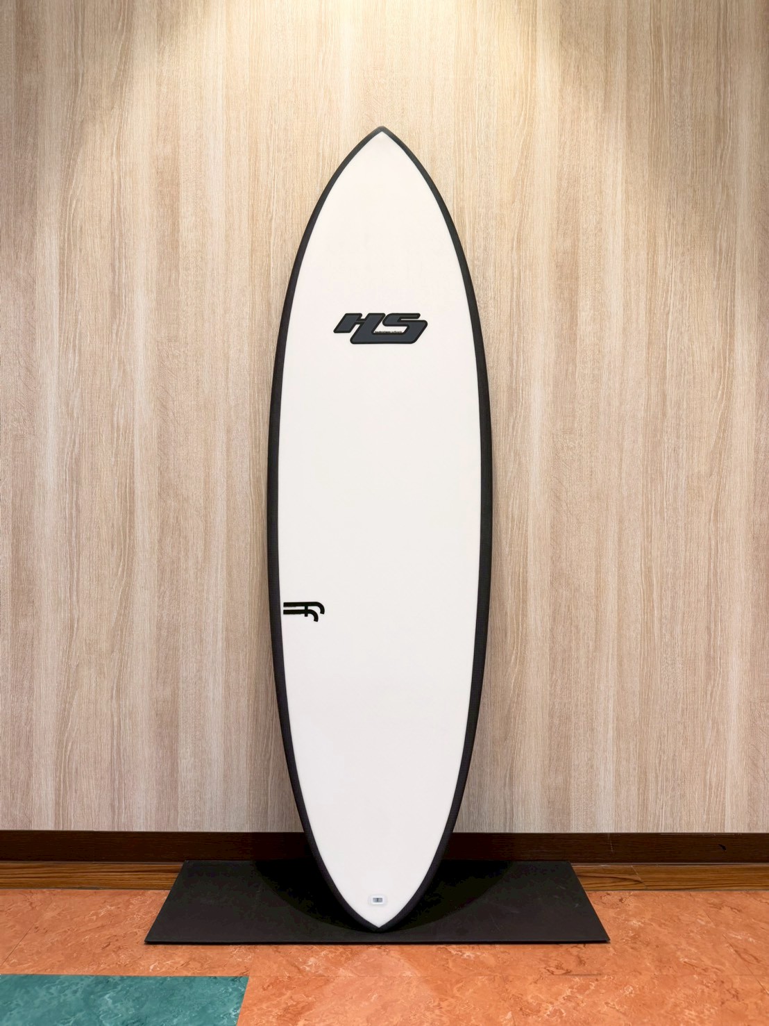 LINE_ALBUM_2026 ヘイデン_260427_2 - SHOP NEWS | ムラサキスポーツオンラインストア 通販 White surfboard with a black edge leaning on a black mat against a wood-paneled wall, featuring an 'HS' logo midboard.