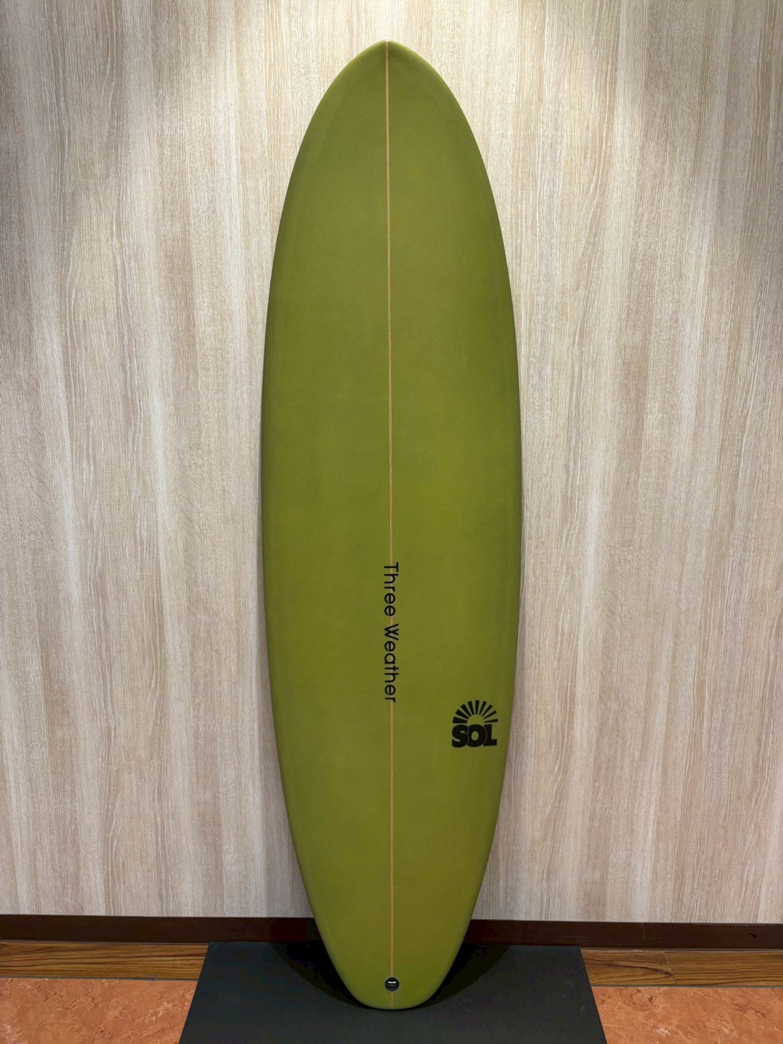 LINE_ALBUM_2026 TW_260422_1 - SHOP NEWS | ムラサキスポーツオンラインストア 通販 Green surfboard standing upright against a wood panel wall, with a thin center rail and SOL logo near the bottom on a black display stand.