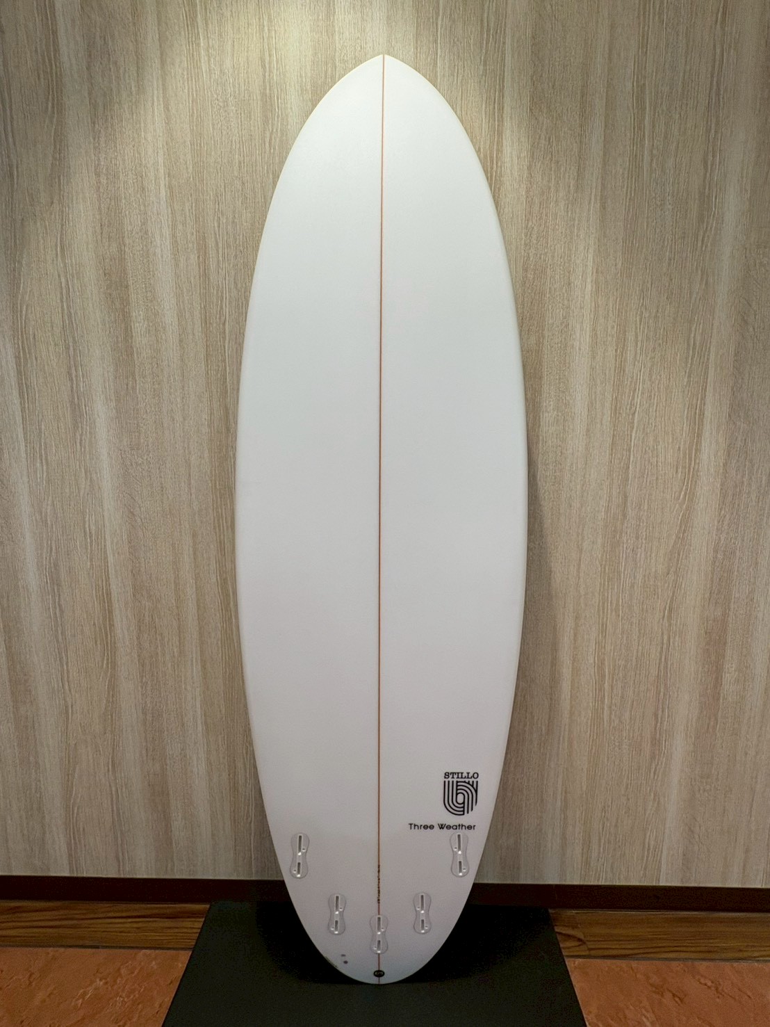 LINE_ALBUM_2026 TW_260422_13 - SHOP NEWS | ムラサキスポーツオンラインストア 通販 White vertical surfboard with an orange center stringer, standing on a black mat against a wood-paneled wall; tail has multiple fin boxes and a small logo near the bottom.