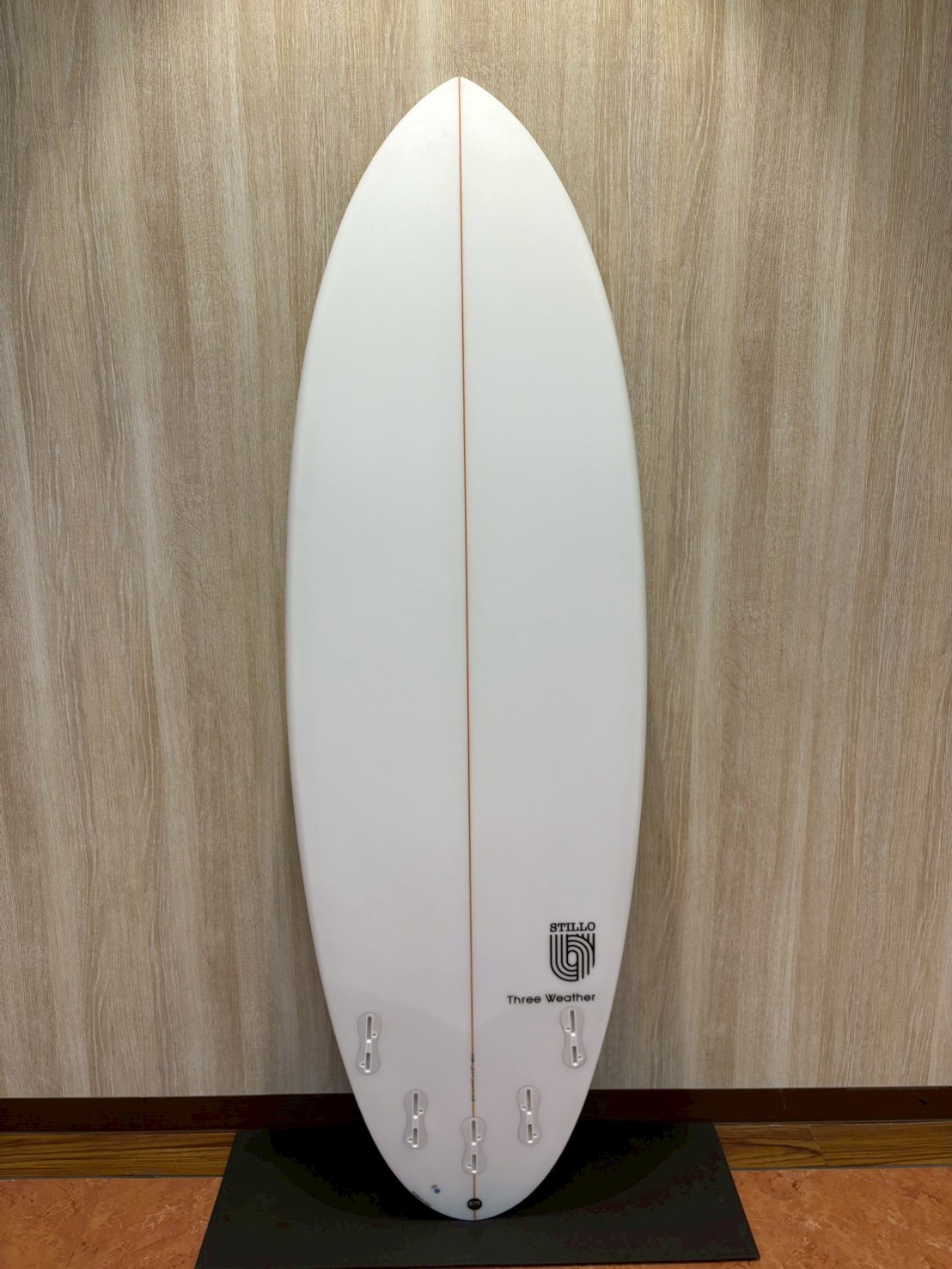 LINE_ALBUM_2026 TW_260422_8 - SHOP NEWS | ムラサキスポーツオンラインストア 通販 White surfboard on a display stand against a wood-textured wall, featuring an orange center stringer and six fin boxes at the tail.