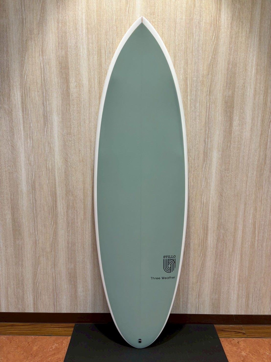 LINE_ALBUM_2026 TW_260423_1 - SHOP NEWS | ムラサキスポーツオンラインストア 通販 Light teal surfboard standing upright against a wooden wall with a white edge and small logo near the bottom that reads 'STILLO' and 'Three Weather'