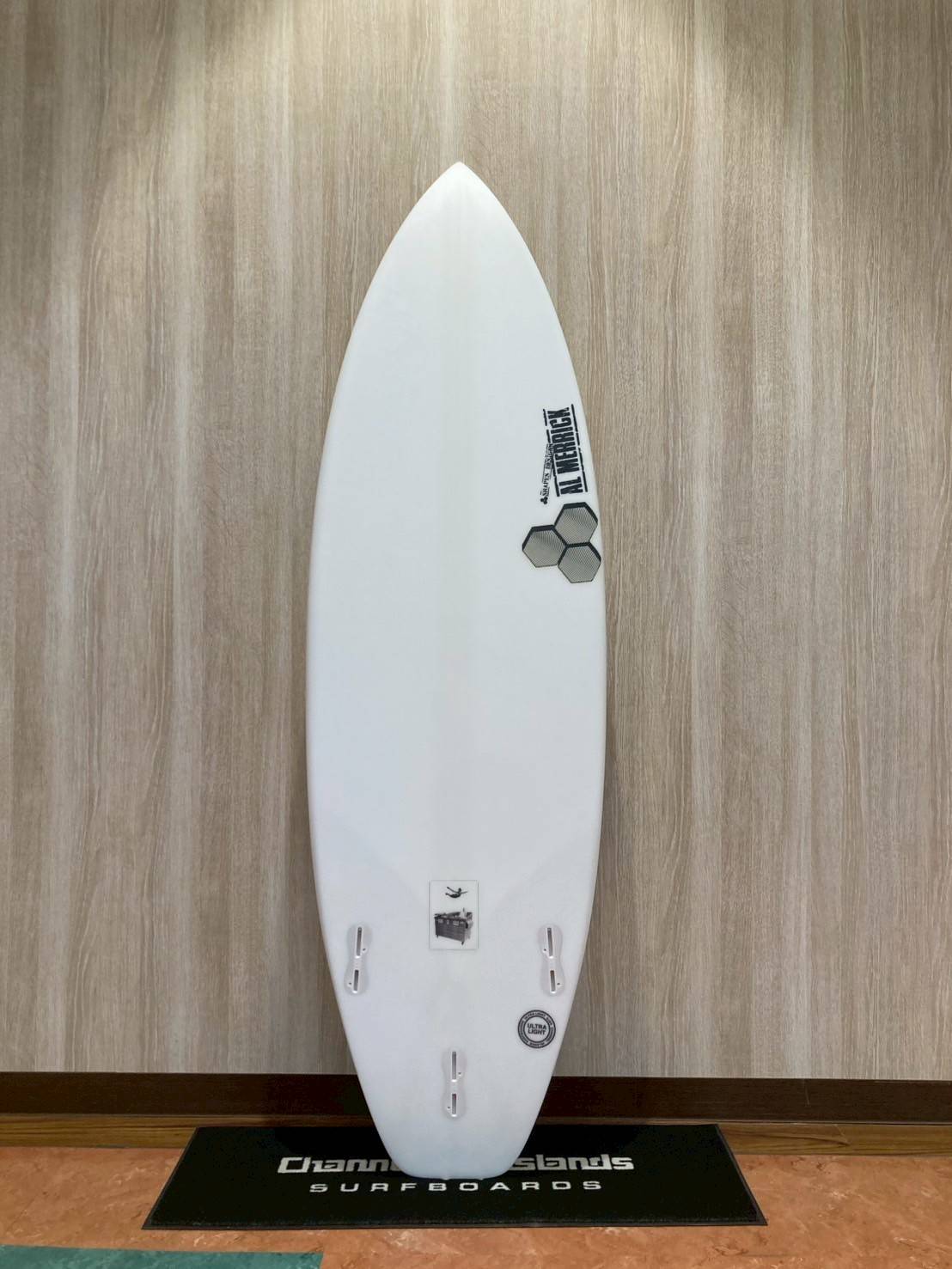 130739 - SHOP NEWS | ムラサキスポーツオンラインストア 通販 White surfboard standing upright against a wood-paneled wall on a display mat with 'Surfboards' branding.