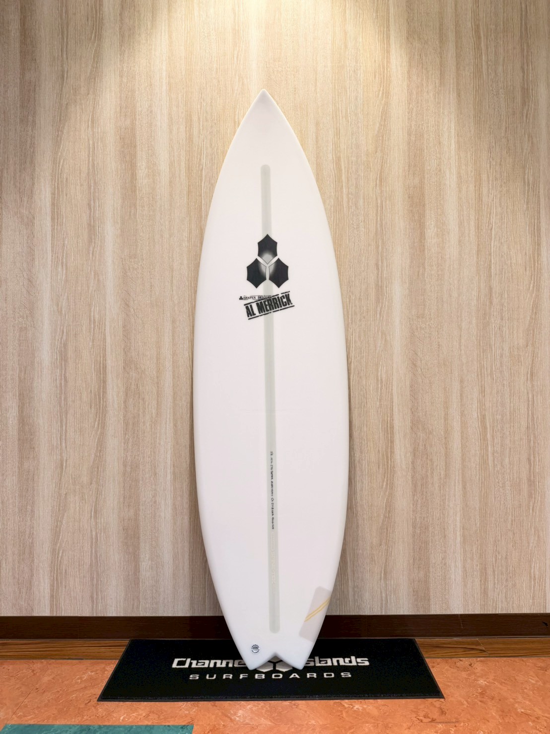 LINE_ALBUM_2026 CI_260427_1 - SHOP NEWS | ムラサキスポーツオンラインストア 通販 White surfboard standing upright against a wood-paneled wall with a branded black mat at its base.
