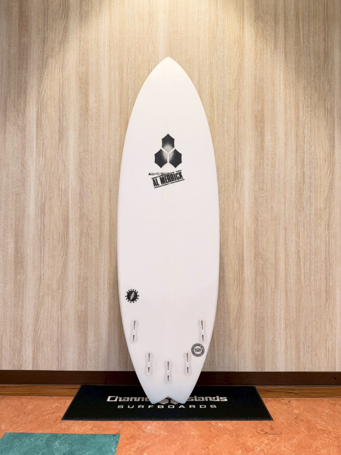LINE_ALBUM_2026 CI_260427_2 - SHOP NEWS | ムラサキスポーツオンラインストア 通販 White surfboard on a display stand against a wood-paneled wall, showing multiple fin boxes and logos (Al Merrick)
