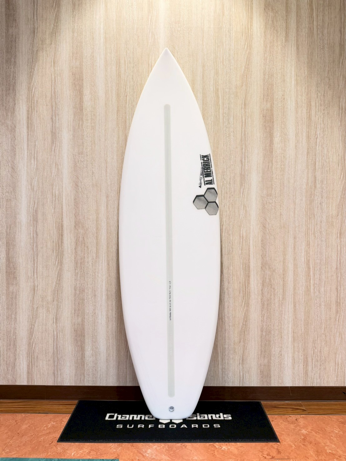 LINE_ALBUM_2026 CI_260427_3 - SHOP NEWS | ムラサキスポーツオンラインストア 通販 White surfboard with a pale gray stringer stands upright on a black display mat against a wood-paneled wall.
