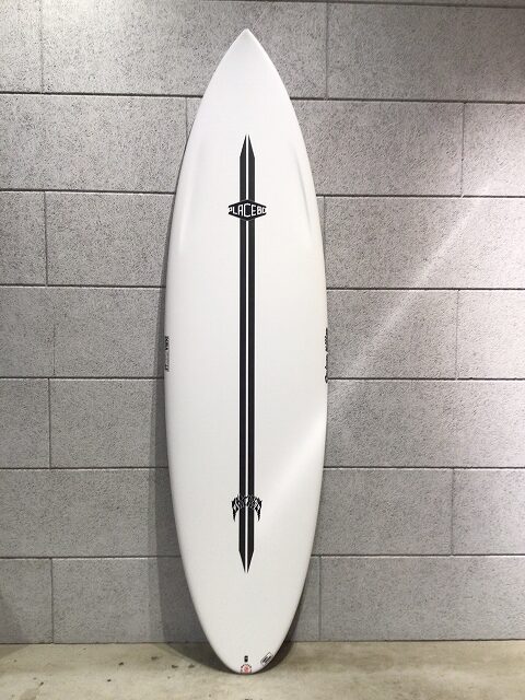 s-image0 - SHOP NEWS | ムラサキスポーツオンラインストア 通販 White surfboard with a black vertical stripe and logos, leaning against a gray tiled wall.