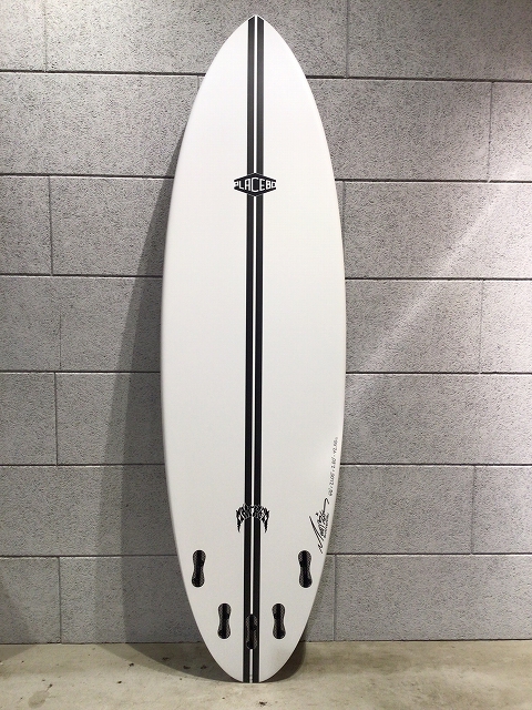 s-image1 - SHOP NEWS | ムラサキスポーツオンラインストア 通販 White surfboard with black center stripes, a small logo, leaning against a gray brick wall near the floor.