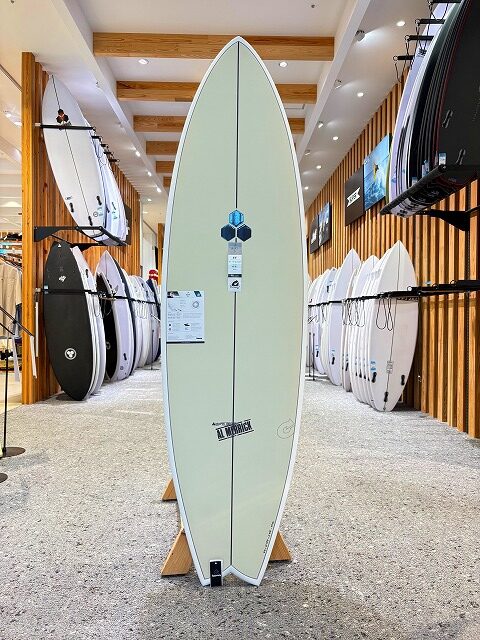Cream-colored surfboard on a display stand inside a surf shop, with other boards visible on racks around it