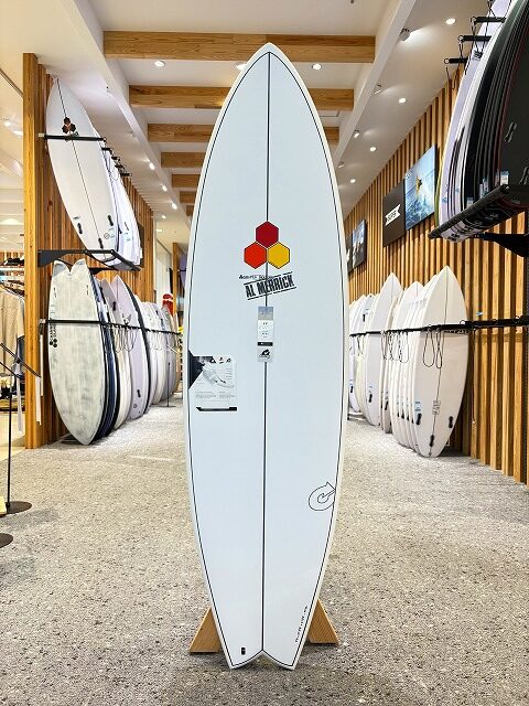 White surfboard on a display stand in a surf shop, centerboard features a red/yellow hex logo and branding; rows of other boards line the walls on both sides.