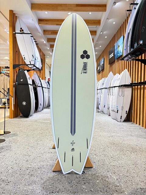 Pale green longboard surfboard with a dark stripe and hexagonal logo, standing on a wooden rack inside a surf shop with other boards on display.