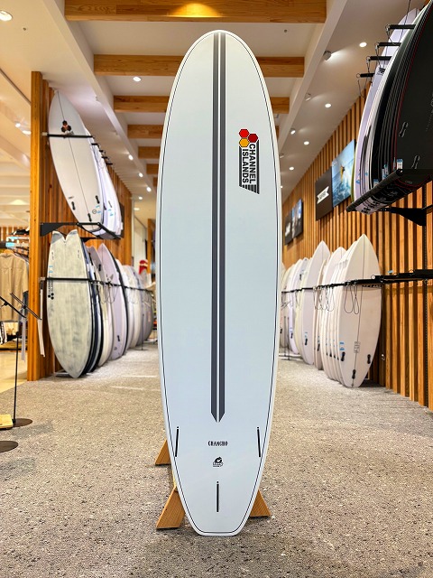 White surfboard on a stand in a surf shop, featuring a vertical gray center stripe and Channel Islands logo near the top.