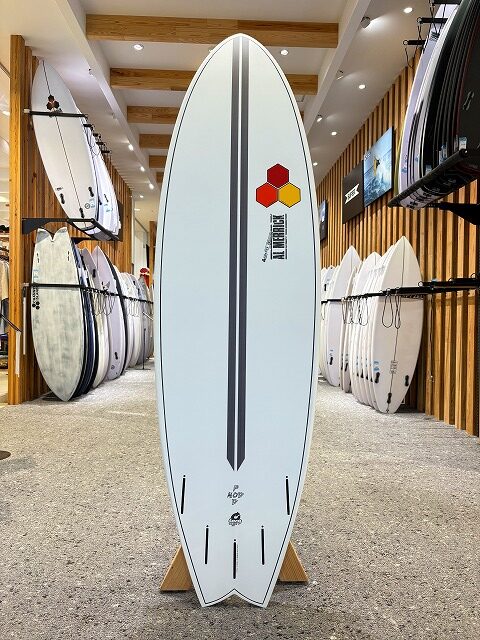 White surfboard on a stand with a dark vertical stripe and hexagon logo, displayed in a surf shop.