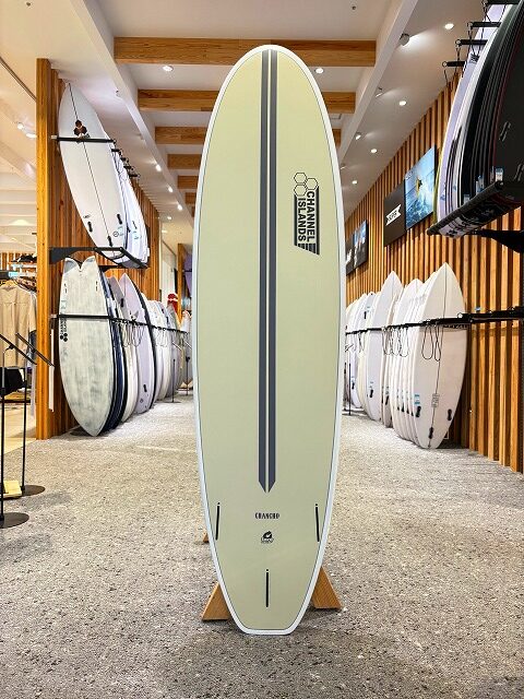 Pale yellow surfboard with a dark vertical stripe on a stand in a surf shop showroom, surrounded by other boards on racks.