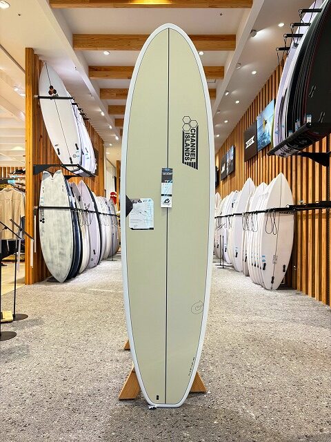 Beige longboard surfboard on a wooden display stand in a surf shop, with rows of boards lining the walls.