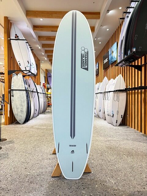 Light blue surfboard with a vertical dark stripe on a wooden stand inside a surf shop, logo visible, surrounded by other boards on racks.