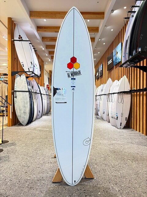 White surfboard on a wooden stand displayed upright in a surf shop, with racks of boards along the walls.