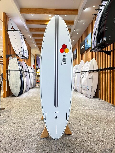 White surfboard on a wooden stand in a surf shop, featuring a black center stripe and logo near the top, with other boards on racks around it.