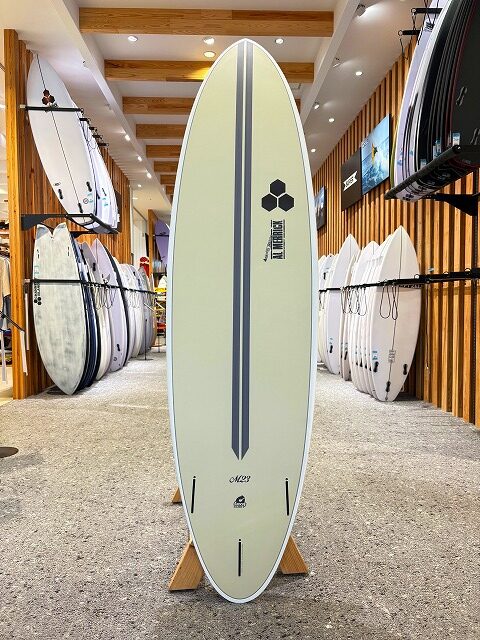 Cream surfboard with dark dual stripes and a central line, upright on a wooden stand in a surf shop, with racks of boards along the walls.