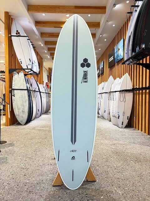 Light blue surfboard on a wooden stand inside a surf shop, with racks of boards lining both sides in the background.