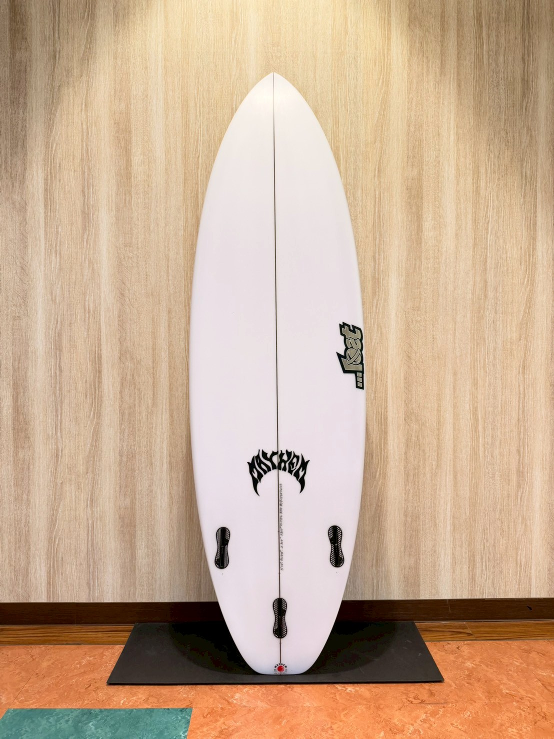 LINE_ALBUM_2026 LOST_260427_2 - SHOP NEWS | ムラサキスポーツオンラインストア 通販 White surfboard standing upright on a black mat, leaned against a wood-paneled wall, with four fin boxes and logos visible.