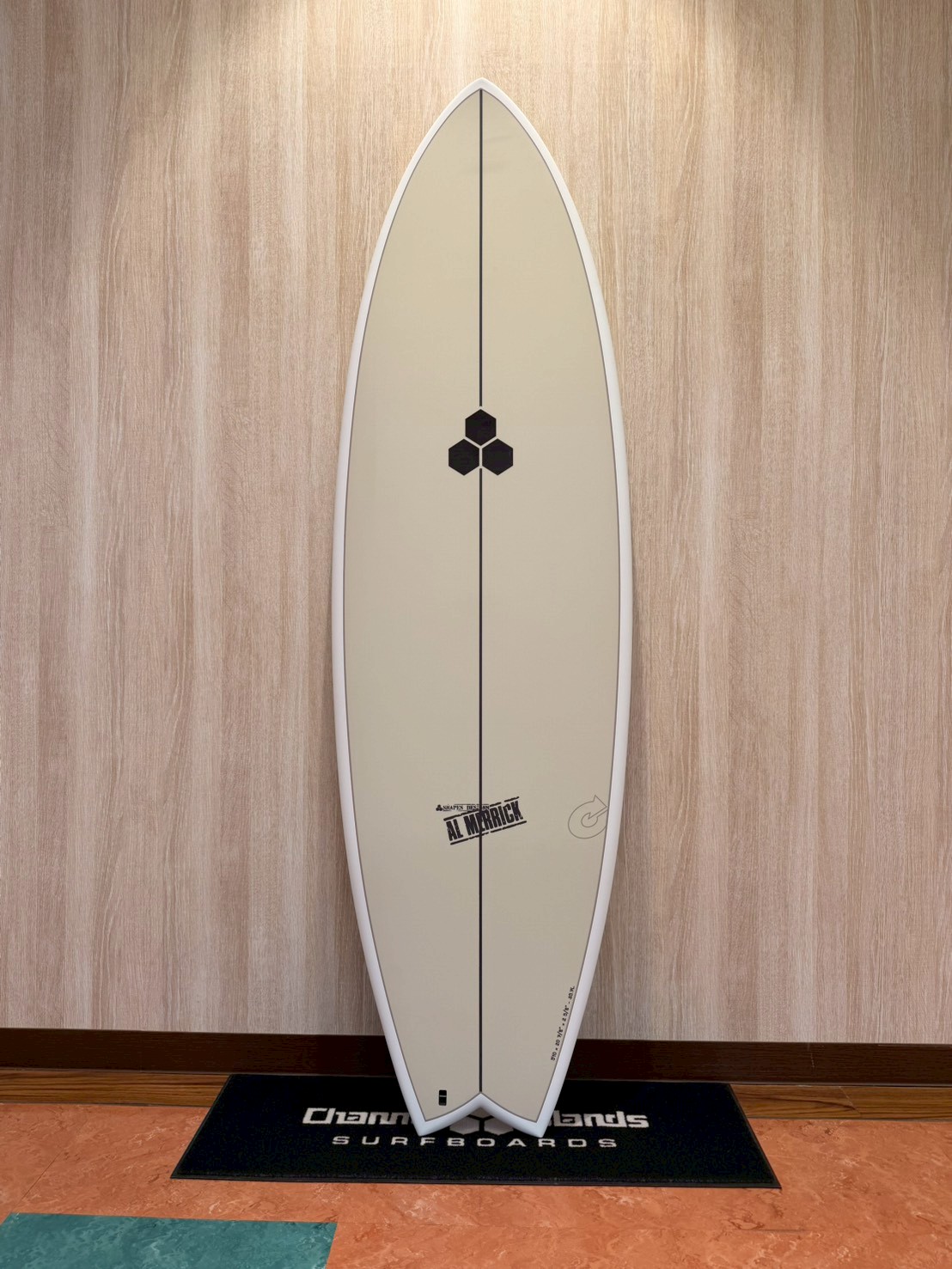 LINE_ALBUM_2026 トルク_260426_1 - SHOP NEWS | ムラサキスポーツオンラインストア 通販 Beige surfboard standing upright against a wood-paneled wall on a display mat, featuring a center black stringer and hexagon logo near the middle.