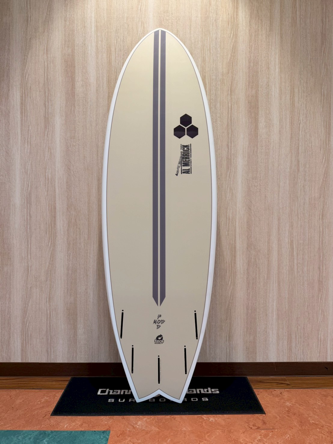 LINE_ALBUM_2026 トルク_260426_2 - SHOP NEWS | ムラサキスポーツオンラインストア 通販 Beige surfboard with two dark vertical stripes and small logos, standing upright on a display mat against a wood panel wall.