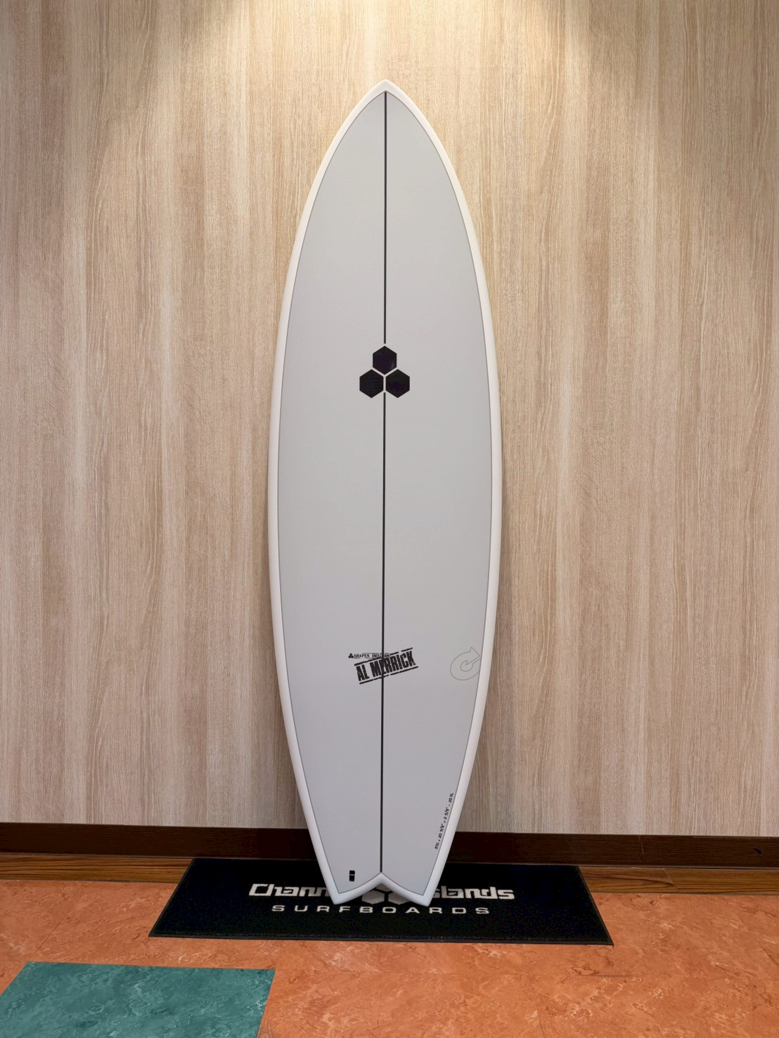 LINE_ALBUM_2026 トルク_260426_5 - SHOP NEWS | ムラサキスポーツオンラインストア 通販 White surfboard on a display stand leaned against a wooden wall, with a black hexagon logo in the center and a vertical stringer, labeled Al Merrick at the bottom edge.