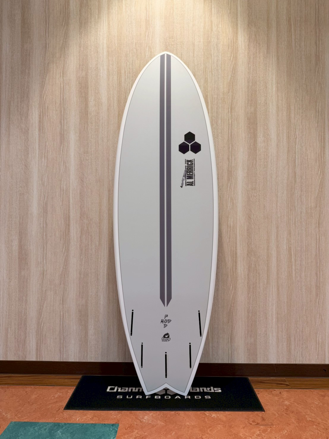 LINE_ALBUM_2026 トルク_260426_6 - SHOP NEWS | ムラサキスポーツオンラインストア 通販 White surfboard standing upright against a wooden wall with a dark gray center stripe and logo near the middle, displayed indoors on a mat.