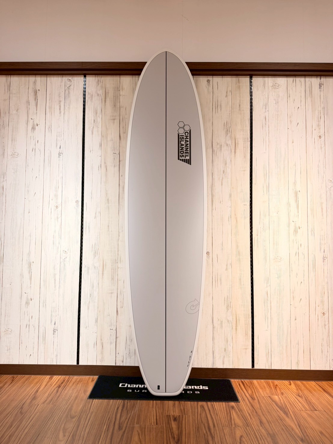 LINE_ALBUM_2026 トルク_260426_7 - SHOP NEWS | ムラサキスポーツオンラインストア 通販 White surfboard standing upright against a light wood-paneled wall, centered on a black Channel Islands mat.