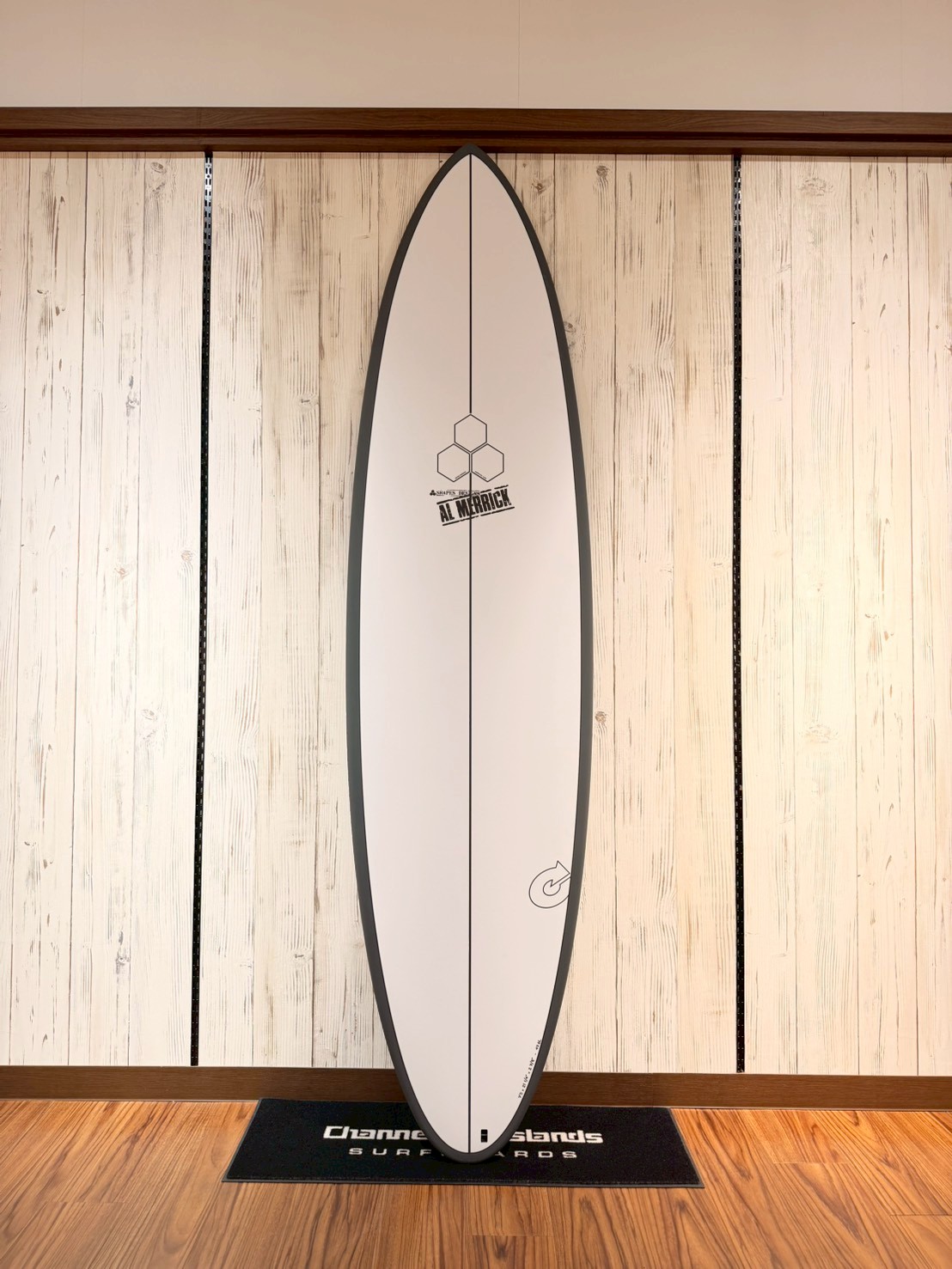 LINE_ALBUM_2026 トルク_260426_9 - SHOP NEWS | ムラサキスポーツオンラインストア 通販 White surfboard with gray rails, standing upright on a wall display in a store, showing a hexagon logo and AL MERRICK text near the center.