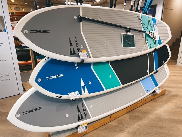 Stack of stand-up paddleboards on a showroom display rack, gray, blue, teal, and black boards with logos visible.