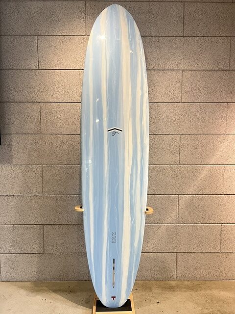 s-IMG_5679 - SHOP NEWS | ムラサキスポーツオンラインストア 通販 Blue-and-white striped surfboard standing upright against a beige brick wall on a wooden rack, ready for display or storage.