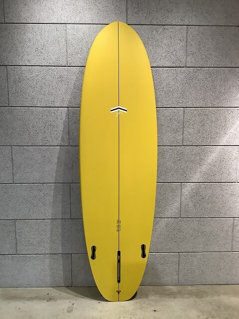 s-IMG_6378 - SHOP NEWS | ムラサキスポーツオンラインストア 通販 Yellow surfboard standing upright against a gray tiled wall, showing two fin boxes and a bottom leash plug near the tail.