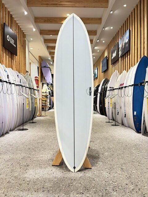 White surfboard on a display stand in a surf shop showroom, with rows of other boards lining the walls and bright overhead lighting.