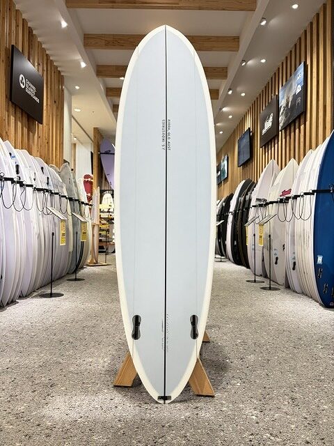 White surfboard on a wooden stand in a surf shop, with rows of boards along the walls behind it.