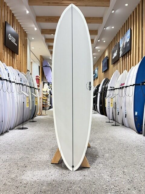 Tall white surfboard standing upright on a wooden display in the center of a surf shop, flanked by rows of other boards on racks.