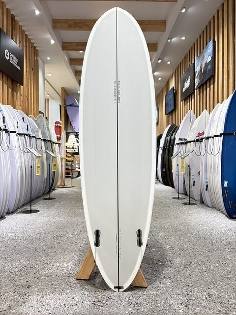 White longboard surfboard on a stand in a showroom, with racks of boards along the walls.