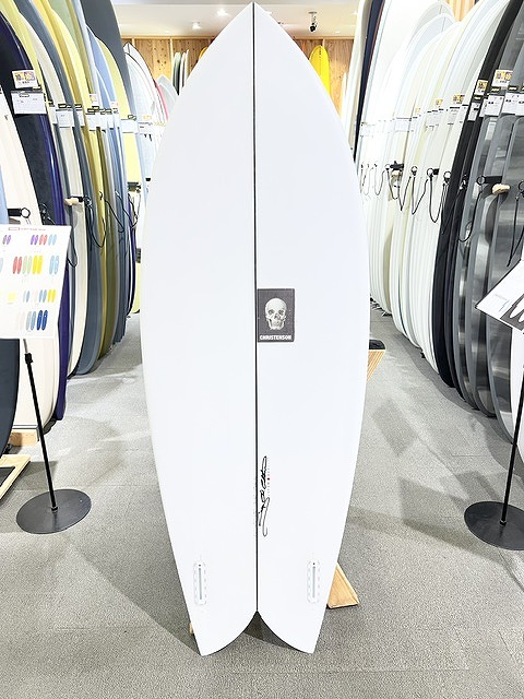 White surfboard with a black skull logo stands upright on display among colorful boards in a surf shop showroom.