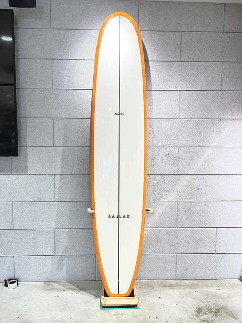 White surfboard with an orange edge standing upright on a wooden stand against a gray tiled wall.