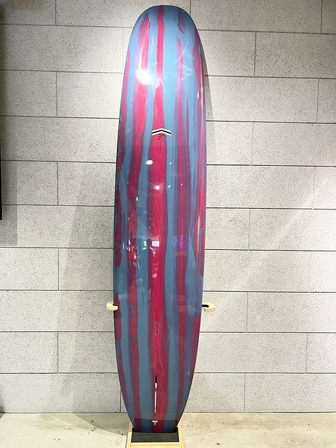 s-IMG_6376 - SHOP NEWS | ムラサキスポーツオンラインストア 通販 Colorful surfboard with pink and blue vertical stripes standing on a wall rack against a gray brick wall, logo visible in center.