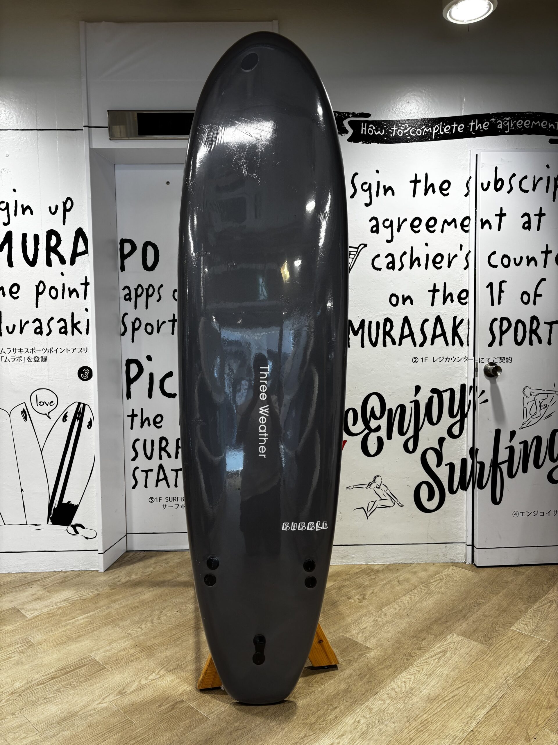 Glossy black surfboard standing upright on a wooden rack indoors, with white walls covered in bold, hand-written surfing text in the background.