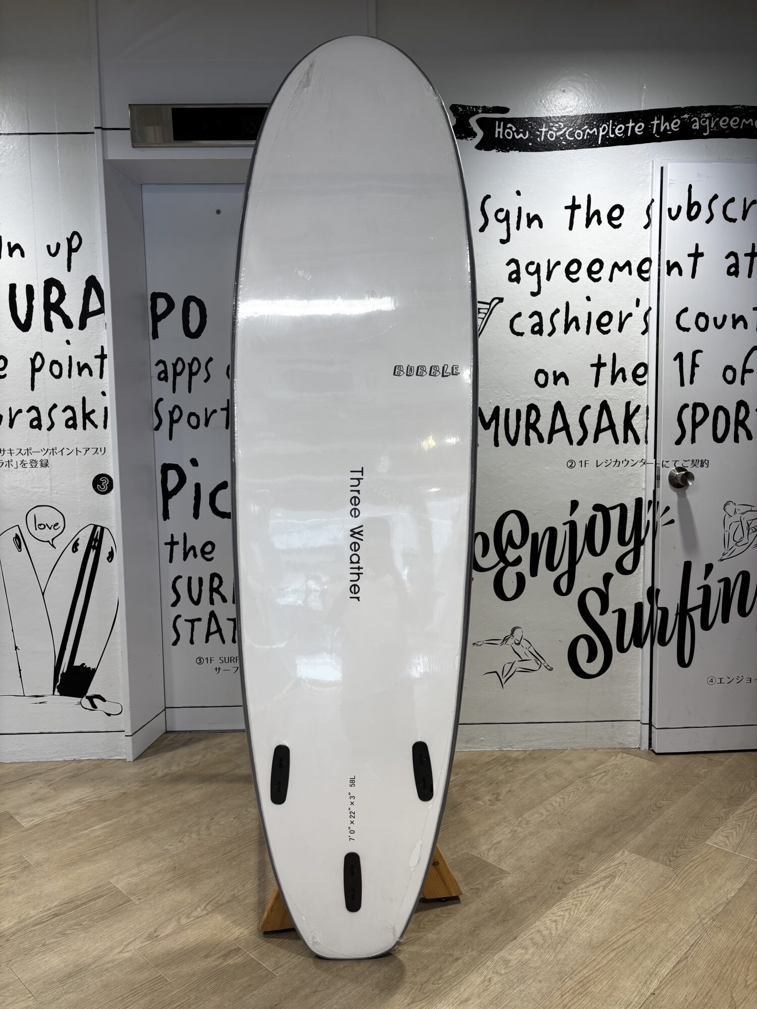 White surfboard standing upright indoors on a wooden floor, with black graffiti-style text and drawings on the walls behind it, and black fin slots visible at the bottom of the board.